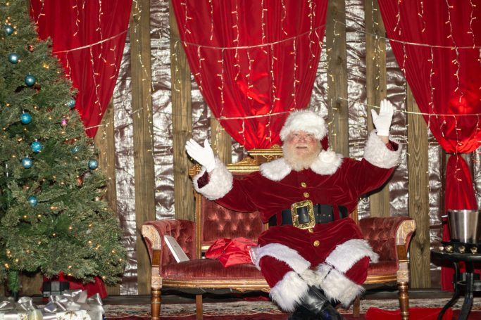 Santa Claus in a red suit sitting on a sofa with a Christmas tree and festive decorations.