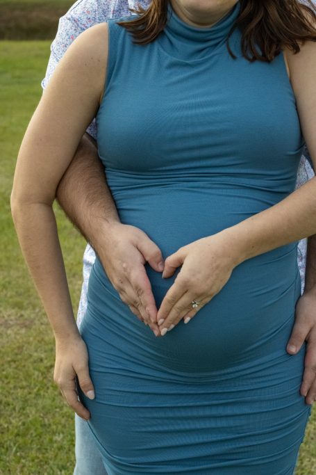 Pregnant woman in a teal dress forming a heart shape with her hands on her belly.